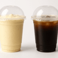 Two clear plastic cups with dome lids - one containing a creamy banana milkshake and the other containing dark cold coffee, both with condensation droplets on a white background